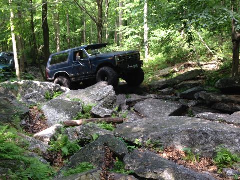 1988 Ford Bronco - A dark blue 1987 Ford Bronco tackles a challenging rocky path in a wooded area, with another vehicle visible in the background.