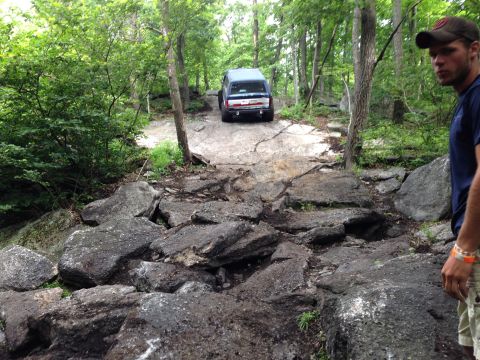 1988 Ford Bronco - A dark SUV tackles a challenging rocky path through a dense forest.