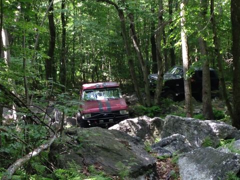1988 Ford Bronco - Two off-road vehicles traverse a dense forest with large rocks.