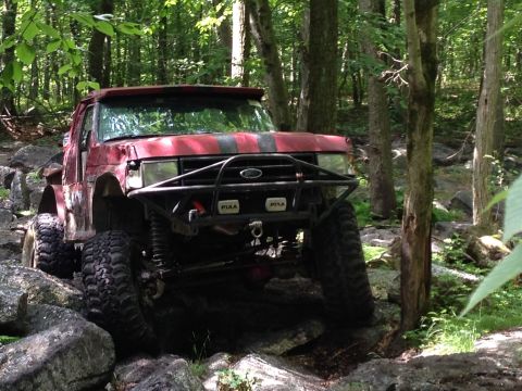 1988 Ford Bronco - This modified truck is shown off-road, with large tires and a reinforced bumper, on a rocky trail surrounded by trees.