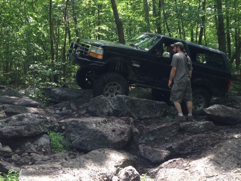 1988 Ford Bronco - A man watches a black Ford Bronco traverse a challenging rocky terrain in a wooded area.