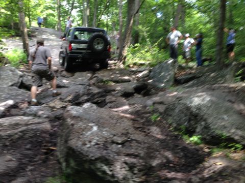 1988 Ford Bronco - A black SUV tackles a challenging rocky terrain, with several onlookers observing the off-road adventure.