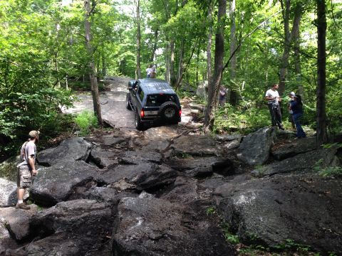 1988 Ford Bronco - An off-road vehicle tackles a challenging rocky path in a wooded area with onlookers observing the action.