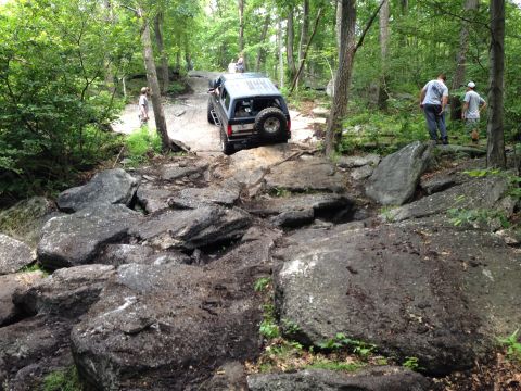 1988 Ford Bronco - An SUV tackles a challenging off-road trail with large rocks and trees surrounding it.