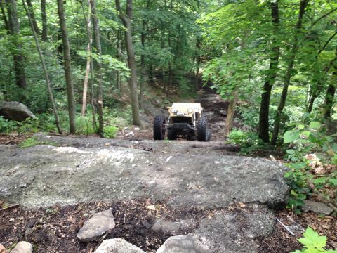 1988 Ford Bronco - A custom-built off-road buggy navigates a challenging, wooded trail with large rocks and mud.