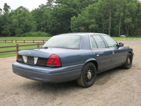 2001 Ford Crown Victoria - A blue 2001 Ford Crown Victoria sedan is parked on a gravel road with trees in the background.