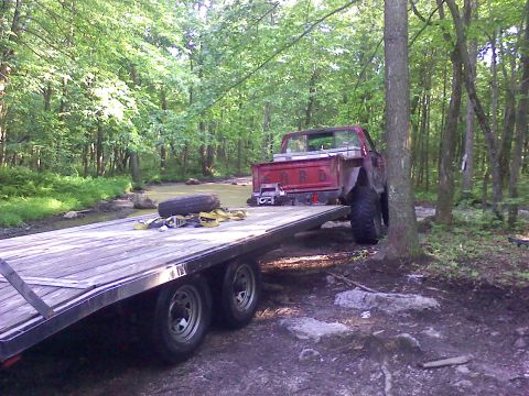 1988 Ford Bronco - A red pickup truck is loaded onto a trailer, parked on a dirt path surrounded by trees and a stream.