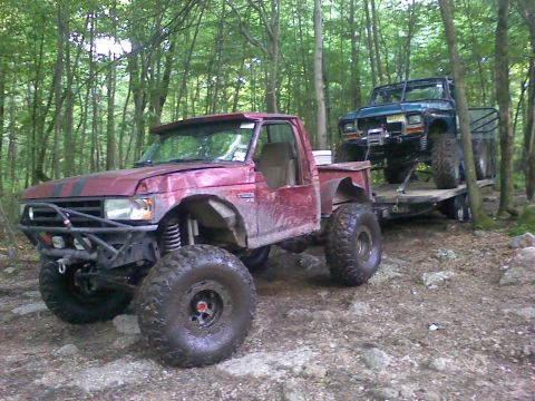 1988 Ford Bronco - Two heavily modified off-road vehicles are seen in a wooded area, one being towed on a trailer.