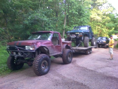 1988 Ford Bronco - A red pickup truck with large off-road tires tows a green SUV on a trailer down a dirt road.