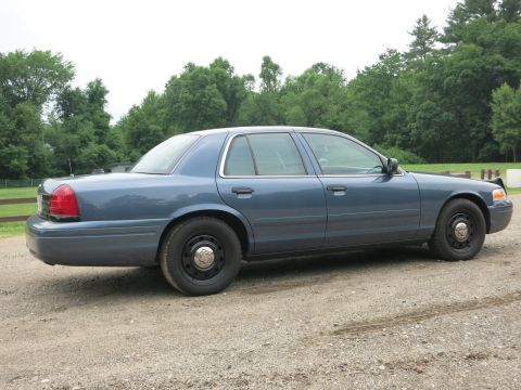2001 Ford Crown Victoria - A blue 2001 Ford Crown Victoria sedan is parked on a gravel surface with trees in the background.