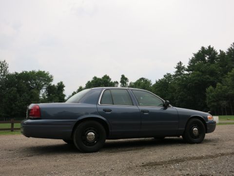 2001 Ford Crown Victoria - A blue 2001 Ford Crown Victoria is parked outdoors on a gravel surface with a wooden fence and trees visible.