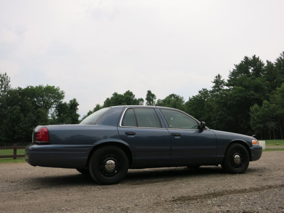 Side view of a blue 2001 Ford Crown Victoria parked on a gravel lot with trees in the background