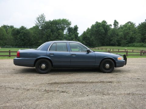 2001 Ford Crown Victoria - A blue 2001 Ford Crown Victoria sedan with a push bar is parked on a gravel surface with trees in the background.