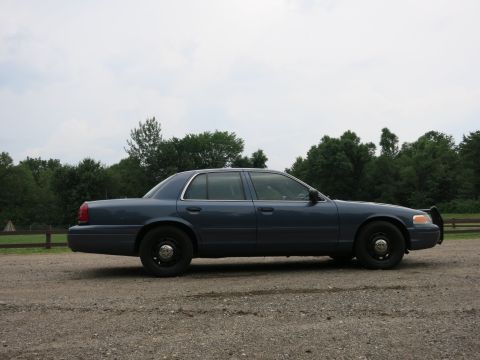 2001 Ford Crown Victoria - Side profile of a blue 2001 Ford Crown Victoria, likely a police vehicle, parked on a gravel surface.