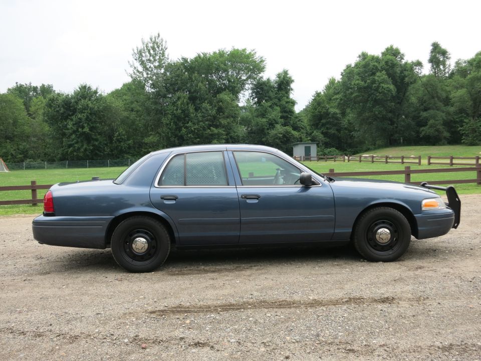 Blue Ford Crown Victoria Police Interceptor parked on a gravel lot with trees in the background