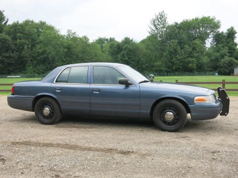 2001 Ford Crown Victoria - A blue 2001 Ford Crown Victoria sedan equipped with a push bar is parked on a gravel surface.