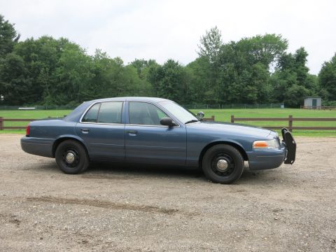 2001 Ford Crown Victoria - A blue 2001 Ford Crown Victoria police car is parked on a gravel lot with trees and a fence in the background.
