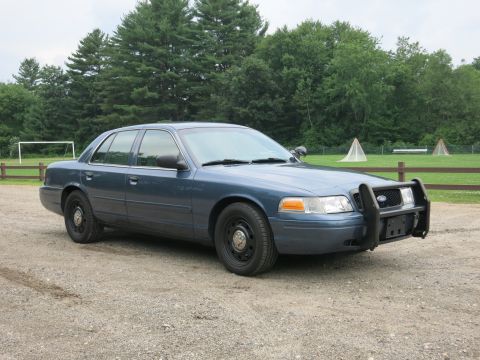 2001 Ford Crown Victoria - A blue 2001 Ford Crown Victoria police car is parked on a gravel lot with trees and a soccer goal in the background.