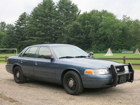 2001 Ford Crown Victoria - A blue 2001 Ford Crown Victoria police car is parked on a gravel surface with trees and a field in the background.