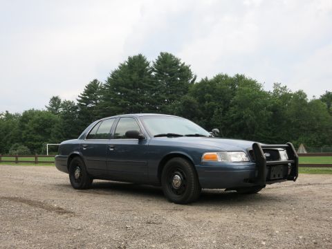 2001 Ford Crown Victoria - A blue 2001 Ford Crown Victoria, equipped with a push bar, is parked outdoors on a gravel surface.