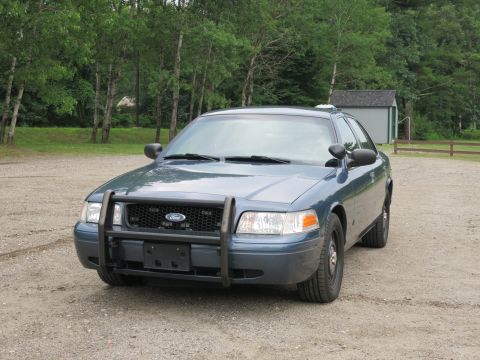 2001 Ford Crown Victoria - A blue Ford Crown Victoria police car is parked outside on a gravel lot, with trees and a small shed visible.
