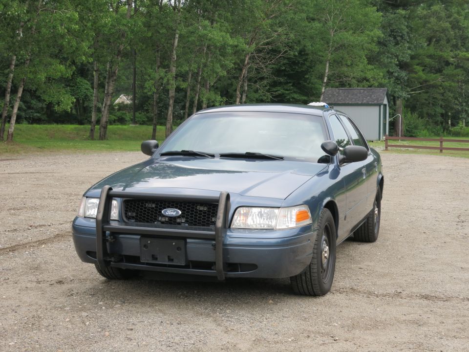 Front view of a blue Ford Crown Victoria police car with a push bumper. It's parked on a gravel lot with trees and a shed in the background