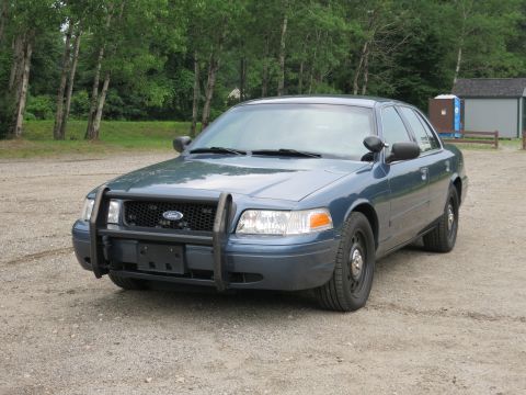 2001 Ford Crown Victoria - A blue Ford Crown Victoria police car is parked on a gravel surface with trees in the background.