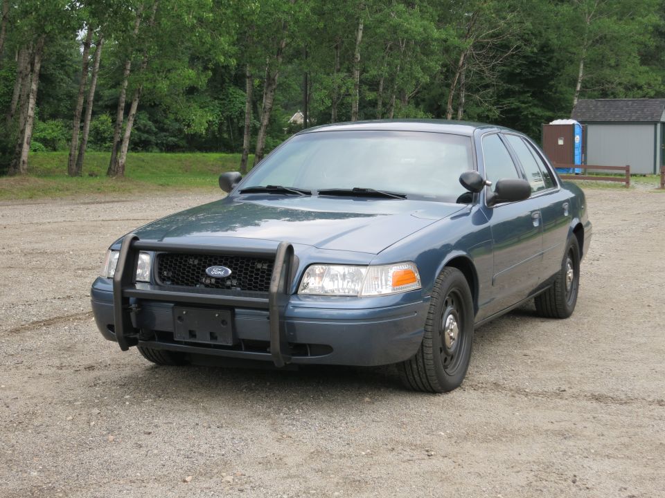 Blue Ford Crown Victoria police car with a push bumper, parked on a gravel lot
