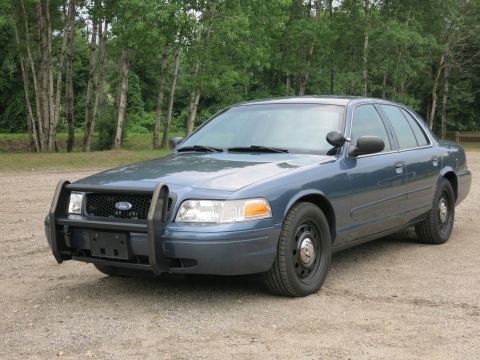 2001 Ford Crown Victoria - A blue 2001 Ford Crown Victoria police car with a push bar is parked on a gravel lot with trees in the background.