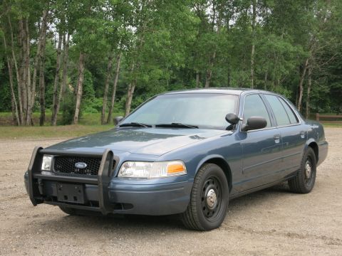 2001 Ford Crown Victoria - A blue Ford Crown Victoria with a push bar is parked outdoors on a gravel surface, with trees in the background.