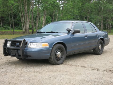 2001 Ford Crown Victoria - A blue 2001 Ford Crown Victoria police car is parked on a gravel surface with trees in the background.