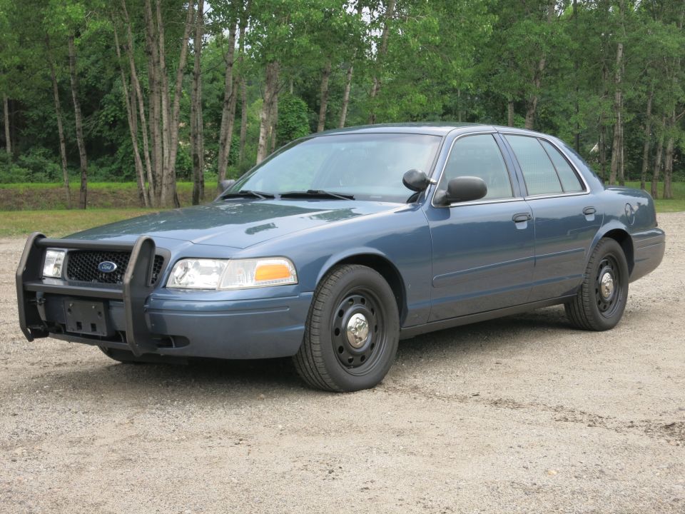 Blue 2001 Ford Crown Victoria police car with a push bumper parked on gravel