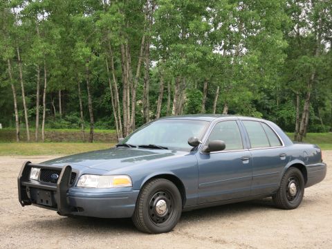 2001 Ford Crown Victoria - A blue 2001 Ford Crown Victoria police car is parked outside with a wooded area in the background.