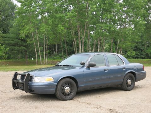 2001 Ford Crown Victoria - A blue 2001 Ford Crown Victoria sedan, equipped with a push bar, is parked outdoors with trees in the background.