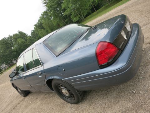 2001 Ford Crown Victoria - A blue 2001 Ford Crown Victoria is shown from a rear three-quarter angle, parked outdoors on a gravel surface with trees in the background.