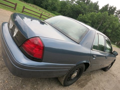 2001 Ford Crown Victoria - The rear quarter panel and taillight of a blue sedan are visible against a backdrop of trees and a wooden fence.