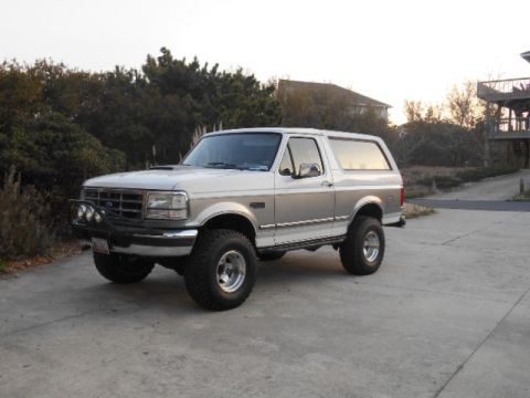 1994 Ford Bronco - This lifted Ford Bronco is parked on a concrete surface with trees and a house in the background.