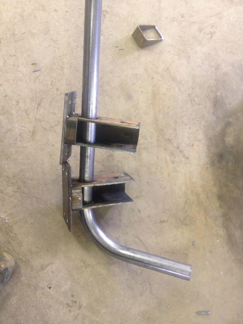 Metal pipe bent and welded to brackets, lying on concrete floor. Small metal square nearby
