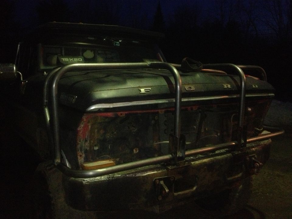 1970 Ford F-250 front end with custom bull bar and missing grille. Dark exterior, red inner fender