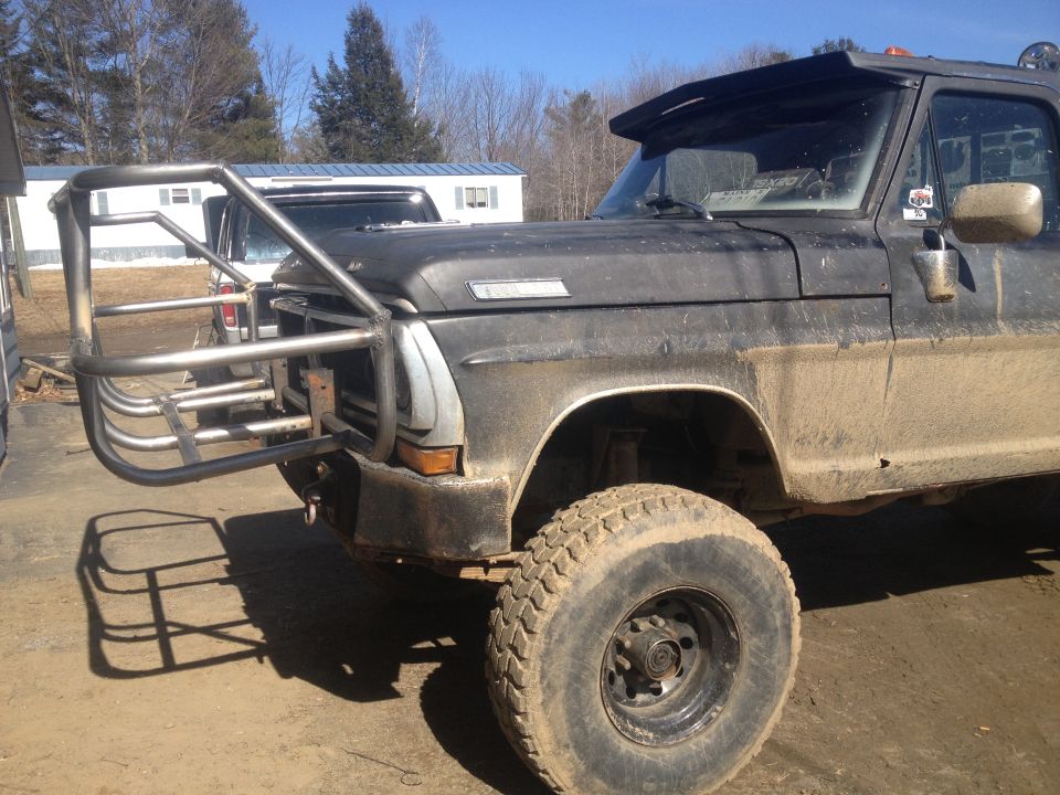 Front quarter view of a dark-colored Ford pickup truck with a large, custom-made metal bumper and brush guard. The truck appears dirty and has large off-road tires