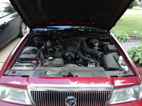 2004 Mercury Grand Marquis - Open engine bay of a maroon Mercury sedan showing the V8 engine, air intake and the Mercury grille emblem below.