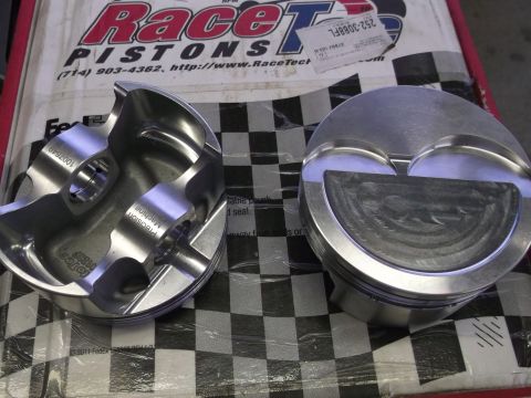 1963 Ford Galaxie - Close-up of two high-performance pistons, likely for a racing engine, displayed on a checkered surface with prominent Race Tech Pistons bra…