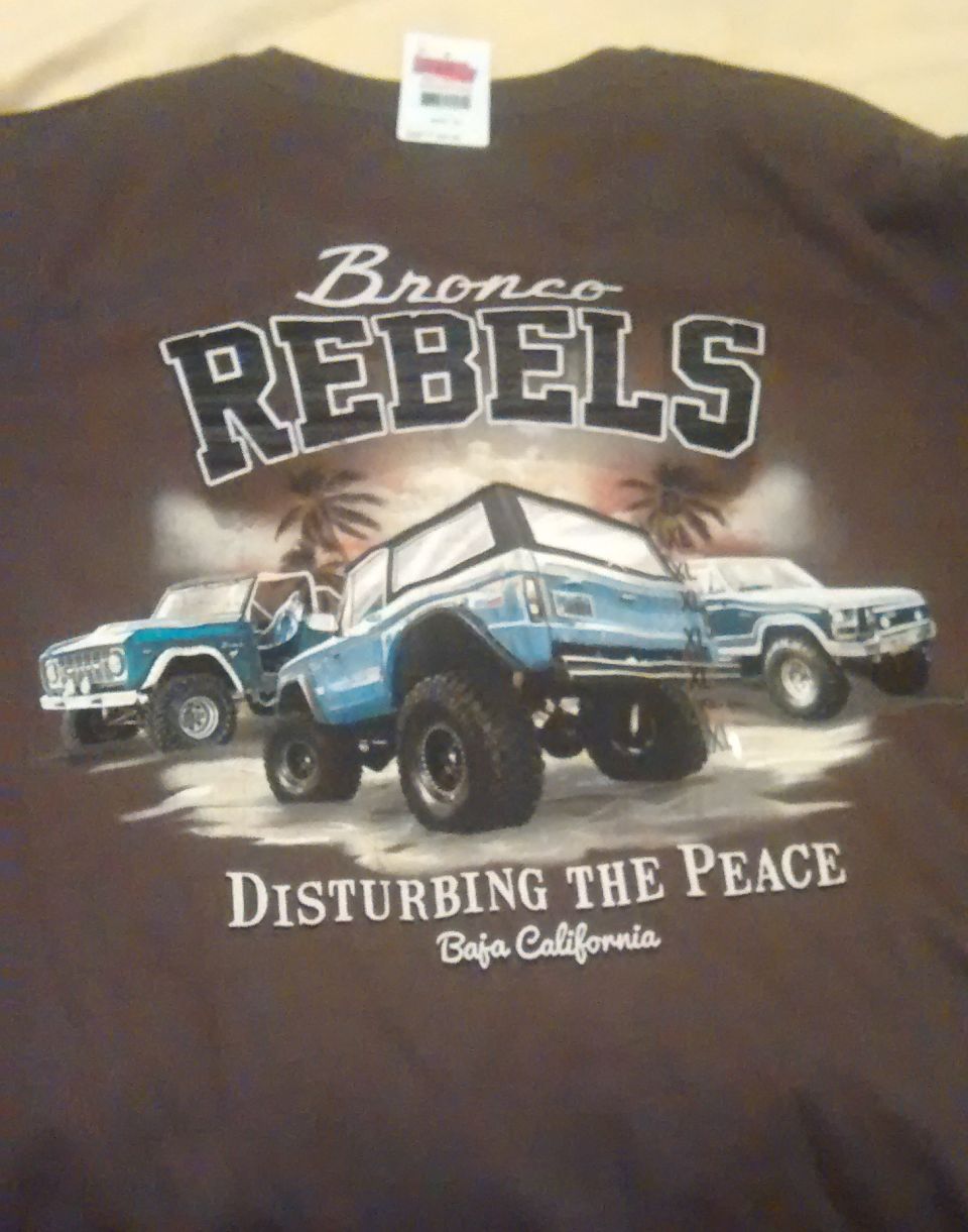 Brown t-shirt graphic showing three Ford Broncos, two in blue and one in white, with palm trees and text 'Bronco Rebels' and 'Disturbing the Peace Baja California'