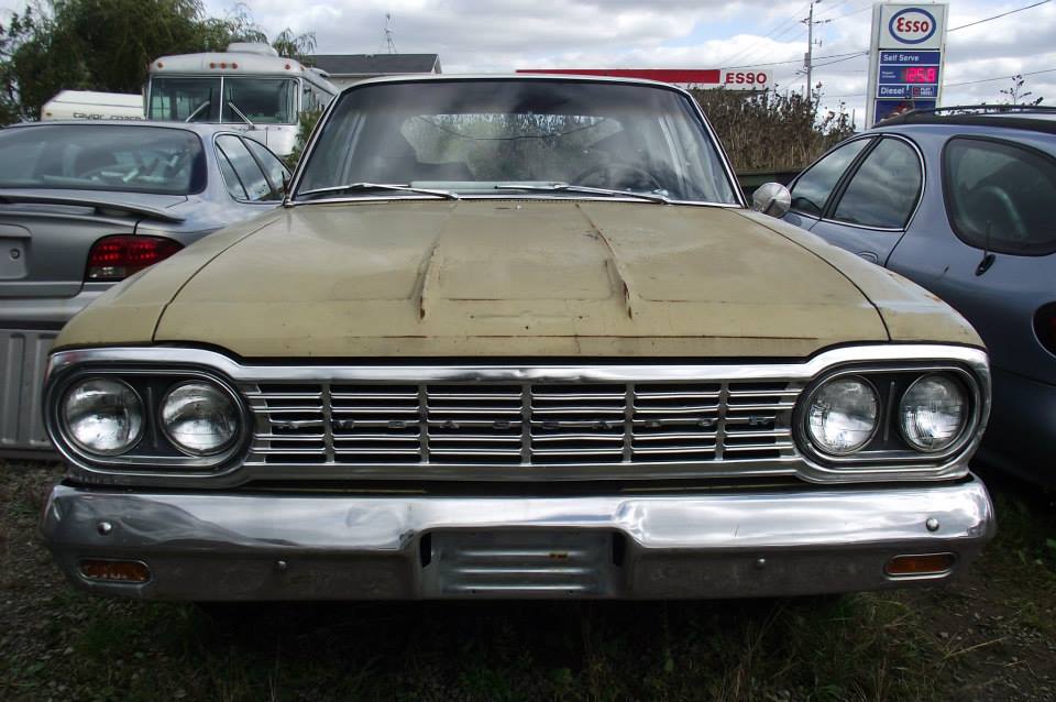 Front view of a tan 1963 American sedan with a chrome grille and dual headlights. Another car and a bus are visible behind it
