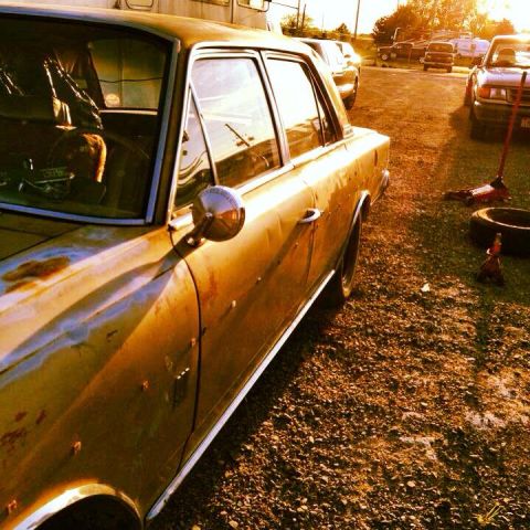 1964 AMC Javelin - A weathered yellow sedan sits in a gravel lot, with other vehicles visible in the background under a bright sun.