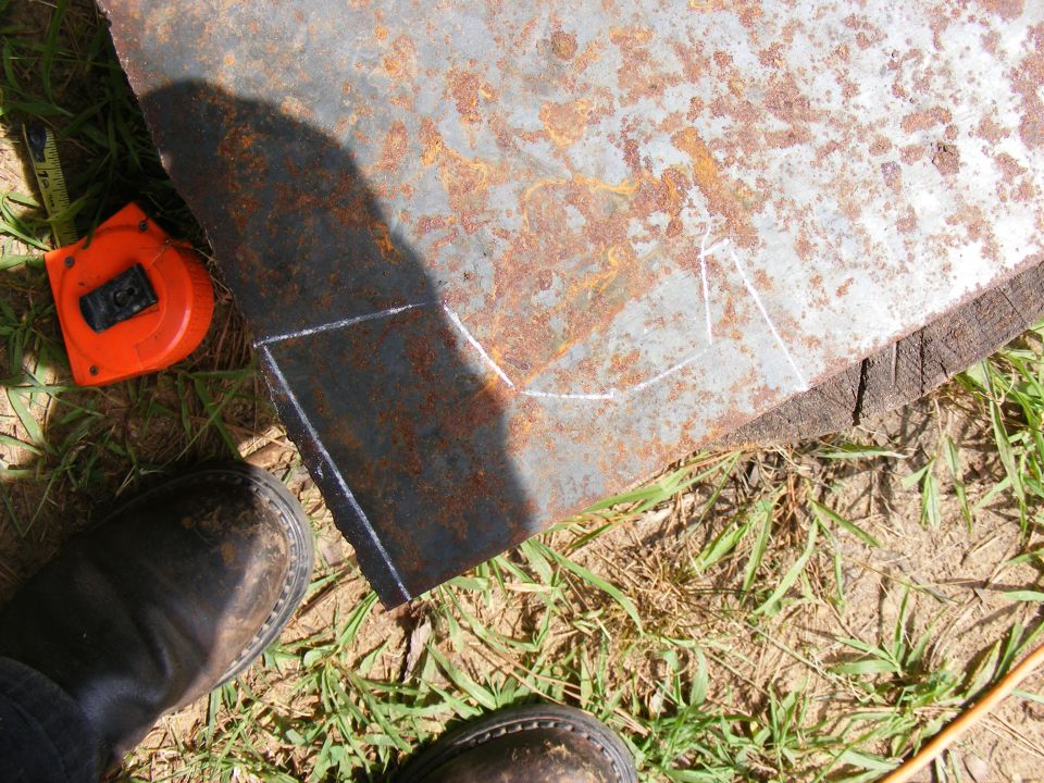 Rusty metal sheet with chalk lines for cutting, orange tape measure, and work boots on the ground