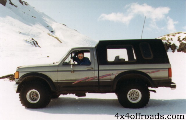 Silver Ford pickup with black camper shell parked in snow, side view, driver visible in window