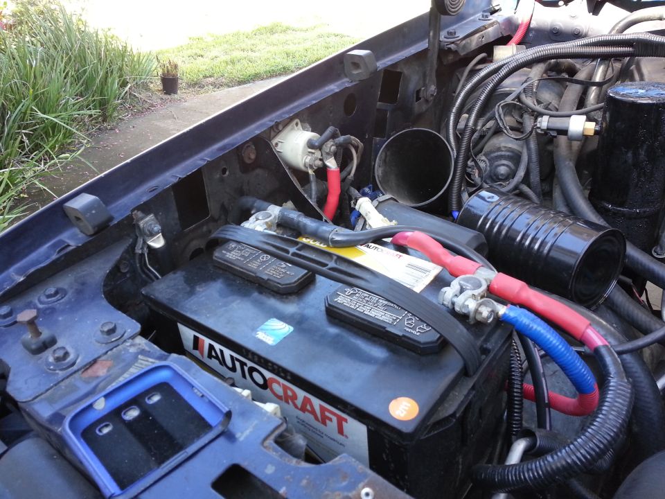 Vehicle engine bay showing a black AutoCraft battery with red and blue cables connected. Some hoses and wiring are visible