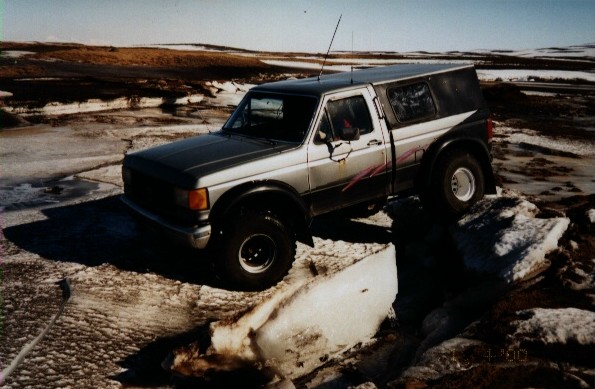 Lifted gray Ford pickup with camper shell crawling over an ice slab in a snowy field