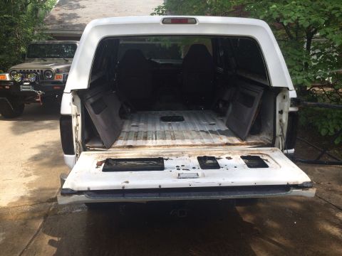 1994 Ford Bronco - The rear of a white SUV is shown with its tailgate down, revealing an empty cargo space. Another vehicle is parked behind it.